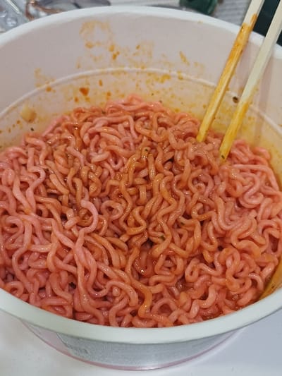 The image displays a close-up view of a white, cylindrical cup containing pink-hued instant ramen noodles. The noodles are curly and coated in an orange-red sauce, with visible splatters on the cup's interior wall. A pair of light-colored chopsticks, showing orange-red residue, are inserted vertically into the noodles. The lower exterior portion of the cup, partially visible, has a faint pink or light purple band. The background is a soft-focused, light-colored surface, indicating an indoor setting. This scene is located within an unspecified indoor location in 서대문구, South Korea. No individuals are visible; the presence of chopsticks suggests the noodles are prepared for consumption.