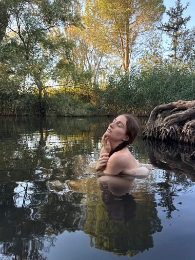 A woman with her eyes closed and head tilted back is in a body of water, possibly a river, with her right arm resting on her chest and her left hand near her face. Her hair is braided. The water is dark and reflective, showing the trees and sky on the opposite bank, along with a large exposed root system on the right side of the image. The surrounding environment is lush with vegetation and trees. The lighting suggests it is either sunrise or sunset. The specific location in Katzrin, Israel cannot be determined from the image alone.