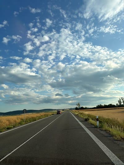 This is a vertical, eye-level, full shot of a paved road stretching into the distance, flanked by fields of dry, golden grass. The sky above is a vibrant blue, dotted with numerous fluffy white clouds that cast shadows on the landscape. A single red car is visible on the road, moving away from the viewer. On the left side of the road, a circular black sign is mounted on a pole, and a white and red striped marker stands on the right. Rolling hills, covered in vegetation and dotted with some trees, are visible in the background. The scene depicts a rural area, likely in Zemplínsky Branč, Slovakia, during daylight.