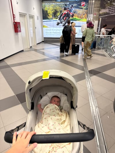 The image is an upward-angle shot of a baby lying in a stroller. The stroller has a light-colored interior with a baby wrapped in a light-colored blanket with pink roses printed on it. Part of the stroller's black handlebar is visible on the right side of the image. In the foreground, a person's hand, wearing a gold ring, holds the stroller's handlebar.

The background reveals an airport terminal setting. Several people are visible, including a person wearing a burgundy hat and a light-green shirt. There are luggage pieces near them. A large advertisement banner for Ducati motorcycles is also present. A red box with a glass panel and a small number "2" on it can be seen. The floor has a patterned gray and white tile design.