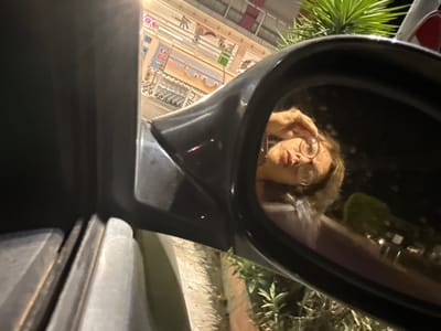 A low-angle, medium shot captures a person's reflection in a car's side mirror. The person, wearing round glasses, is looking into the mirror and adjusting their glasses with their right hand. The background in the reflection shows a brightly lit building with signs, indicating a commercial area. One sign, partially visible, reads "Exit" in English and "Выход" in Cyrillic script, suggesting a location in Cyprus. The car's door and frame are visible in the foreground, along with some foliage on the right side of the frame. The lighting suggests it is evening or night.