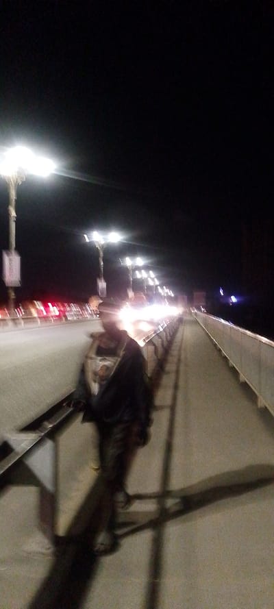 The image depicts a nocturnal scene on a bridge or elevated roadway in Junda ward, Kenya. A single individual, heavily blurred, is positioned in the lower-center foreground, walking on a pedestrian walkway. This person appears to be wearing a dark outer garment, possibly a jacket, over a lighter-colored top. A distinct shadow extends from the person to their right on the concrete surface. To the left of the pedestrian path, multiple vehicle lanes are illuminated by streaks of bright white headlights from oncoming traffic and red taillights from receding vehicles. A series of bright streetlights on tall poles line the left side of the road, providing ambient illumination. A metal guardrail or barrier separates the pedestrian walkway from the vehicle lanes on both sides. The sky is dark and clear, indicating nighttime conditions. The overall image quality is low, characterized by motion blur and noise due to insufficient light.