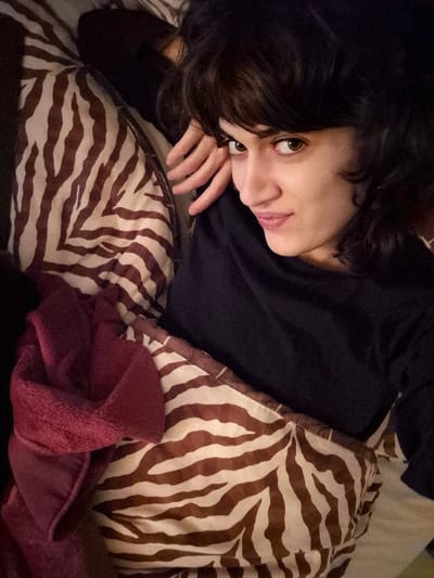 An indoor scene, specified as being in Châtelaillon-Plage, France, depicts a human female positioned horizontally, likely on a bed. The subject, a young adult, has dark, wavy hair and light-colored eyes, and is looking directly at the camera with a slight head tilt and a faint smile. She is wearing a dark, long-sleeved shirt. Her left arm is bent, with the hand partially visible near her head. She is partially covered by bedding featuring a distinct brown and off-white zebra stripe pattern. A folded, maroon-colored, plush blanket is situated on the lower left of the frame, resting on the patterned bedding. The background is indistinct, indicating an interior environment.