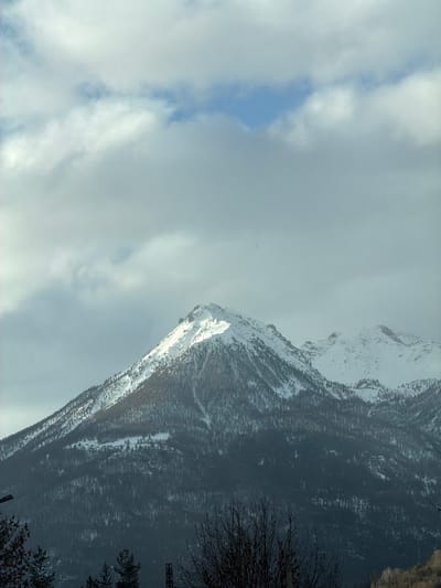 The image shows a mountain partially covered in snow, likely in the vicinity of Briançon, France, given the prompt's context. The sky is overcast with thick clouds, though a small patch of blue sky is visible. The mountain is densely forested, with trees appearing darker against the snow-covered slopes. Some bare tree branches are visible in the lower right corner of the image. The perspective suggests the photograph was taken from a lower elevation looking up at the mountain.