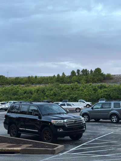 A black Toyota Land Cruiser is parked in a lot in Maryland Heights, Missouri, United States of America. The vehicle is facing left and slightly towards the viewer. The sky is overcast. Behind the Land Cruiser, other vehicles are parked, including a gray SUV, a white pickup truck, and several sedans. A row of trees and vegetation borders a highway embankment in the background. The parking lot is paved with asphalt and marked with white lines.