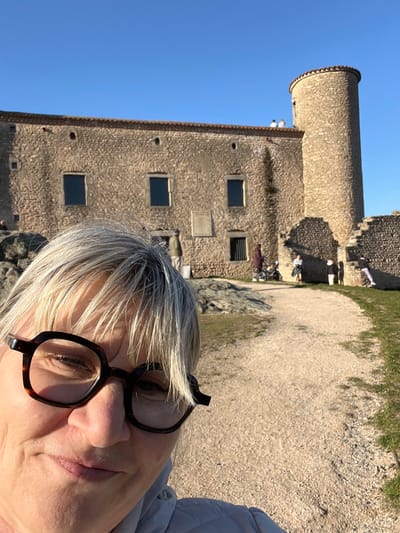 The image depicts an outdoor scene at the Château de Chambles in Chambles, France, under a clear blue sky. In the immediate foreground, centered slightly to the left, is a close-up selfie of a middle-aged individual with light blonde and grey hair, wearing dark-rimmed hexagonal glasses and a light-colored jacket. The individual is looking directly at the camera with a subtle smile.
Behind the foreground subject, the main structure is a large, two-story stone building with a distinctive round tower on its right side. The building features multiple rectangular window openings with dark panes. The roofline has a red tile edge, and two small circular openings are visible higher on the main wall. A light-colored plaque is affixed to the stone wall between the lower-right windows. The lower portion of the tower shows signs of partial ruin.
Several individuals are visible in the mid-ground around the building. Two people are standing on the roof near the tower. On the ground level, to the left of the building, a person is partially visible near a rocky outcrop. Another person in a blue hat is standing near the building's central section. Further right, a person in a dark coat stands near a baby carriage or stroller, with another small group of people nearby. To the far right, a child in purple pants is observed climbing the partially ruined stone wall.
The ground consists of a light-colored, gravel-like path that curves from the lower right towards the center-left of the mid-ground. Areas of green grass and some rocky formations are also present. The scene is illuminated by natural sunlight, casting subtle shadows.