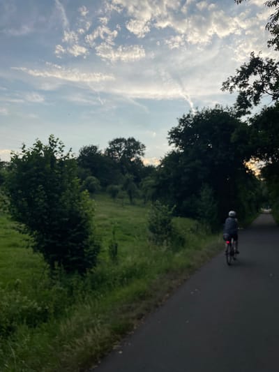 The image captures a cyclist on a paved path in Neukirchen, Germany, amidst a natural, pastoral setting. The cyclist, viewed from behind, is wearing a helmet and dark clothing, and is positioned on the right side of the frame, riding away from the viewer. Their bicycle's red taillight is illuminated. The path curves gently to the right, bordered by lush green grass and vegetation on the left. Beyond the grassy area, a gentle slope rises, dotted with scattered trees and shrubs, suggesting an open field or parkland. The background is dominated by a dense line of dark green trees, creating a natural treeline that extends across the mid-ground. The sky above is a pale blue, partially obscured by wispy white clouds with streaks of contrails, indicating a daytime scene, possibly late afternoon or early evening due to the soft lighting. The overall atmosphere is serene and suggests outdoor recreation in a rural or semi-rural area within or on the outskirts of Neukirchen.