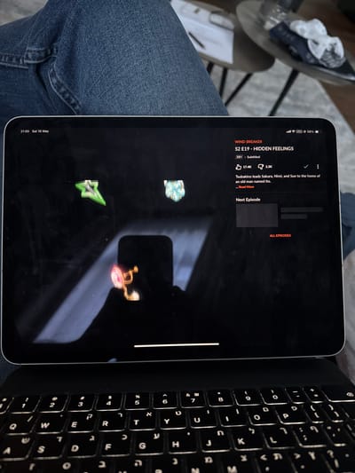The image shows a black iPad Pro, displaying an episode of "Wind Breaker," titled "S2 E19 - Hidden Feelings." The iPad is open on a desk with a keyboard in the foreground. The time is 21:00 and the date is Saturday, 10 May. The episode has been watched 144 times and has 17.4K likes and 2.2K dislikes. The episode description reads: "Tsubakino leads Sakura, Nirel, and Suo to the home of an old man named Itoo." The episode is subtitled. Three icons in the top left corner of the screen are highlighted: a green arrow, a blue flower, and a pink and gold locket. The user can click "Next Episode" or "All Episodes." The coordinates (50.395, 5.93) correspond to a location in Stavelot, Belgium. The specific location cannot be determined from the image.
