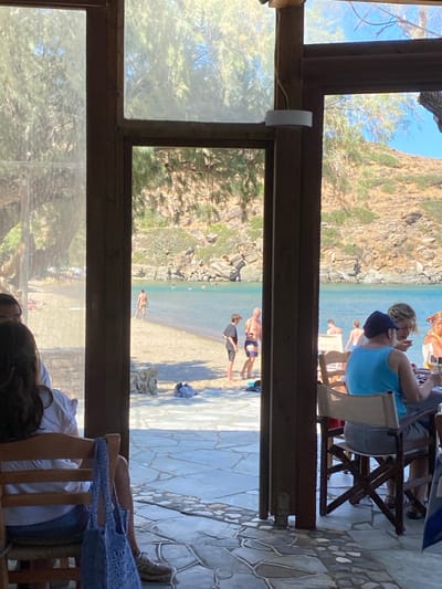 The image captures a sunlit coastal scene in Φάρος, Greece, viewed from inside a structure with a visible wooden framework and a patterned stone-tiled floor. In the foreground on the left, a person with dark hair is seated from behind in a wooden chair, with a light blue woven bag hanging from its back. On the right, multiple individuals are seated at a wooden table, one person wearing a light blue tank top and a dark cap is partially visible. A white cylindrical light fixture is mounted on a central vertical wooden beam.
Beyond the structure, a sandy beach transitions to a calm, clear blue-green body of water. Several individuals are present on the beach: a bare-chested male stands near the water's edge on the left, while two other males, one younger in a dark shirt and one older in striped swim trunks, stand closer to the center. Scattered items, possibly clothing or towels, lie on the sand. Numerous figures are visible swimming or wading in the water. The background features arid, rocky hills with sparse green vegetation, extending down to the coastline under a clear, bright sky.