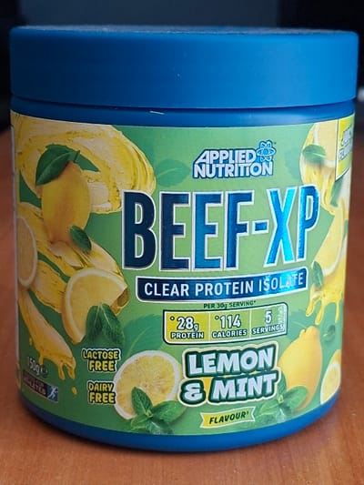 The scene depicts a single cylindrical product container with a blue screw-top lid, positioned upright on a light brown, wood-grain surface. The container's label is predominantly lime green, featuring illustrations of whole lemons, sliced lemons, and fresh mint leaves, with yellow liquid splashes and drips. The brand "APPLIED NUTRITION" is visible at the top center of the label, above the large, metallic blue text "BEEF-XP" which is further described as "CLEAR PROTEIN ISOLATE." Nutritional information indicates "28g PROTEIN," "114 CALORIES," and "5 SERVINGS" per 30g serving. The product's flavor is "LEMON & MINT." Additional text states "LACTOSE FREE," "DAIRY FREE," and "150g e." A small icon of a running figure accompanies "TESTED FOR ATHLETES." The environment suggests an indoor setting, possibly a table or countertop. There are no individuals, actions, or interactions depicted. The location is in the city of Selyanin, Bulgaria.