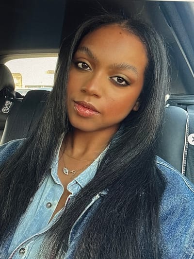 A woman with long black hair and brown eyes looks at the camera, her face angled. She wears a denim shirt, partially unbuttoned to reveal a delicate gold necklace with an eye-shaped pendant. The seat of a car is behind her, visible in the background. She has lipstick, blush, and eyeshadow on her face.