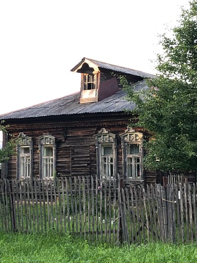The image shows a weathered wooden house with ornate window frames, partially obscured by a wooden picket fence and lush green grass. A tree with dense foliage is visible to the right of the house. The house features a dormer window in the steeply pitched, corrugated roof. The logs of the house are dark and appear aged. The fence is constructed of vertical wooden pickets of varying heights and widths, some leaning or broken. The overall scene suggests a rural or semi-rural setting, consistent with the exterior of a dwelling in Makeikha, Russia. There are no people or animals visible in the image.