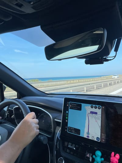 The image captures an interior view from a vehicle in motion on a multi-lane coastal highway in Tirat HaCarmel, Israel. The perspective is forward through the windshield, revealing the Mediterranean Sea bordering the highway under a clear blue sky.
The driver's hands, with light skin tone, are positioned on a black steering wheel on the left side of the frame. The digital instrument cluster visible through the steering wheel indicates a speed of '97' km/h. To the right of the steering wheel, a large, vertically oriented touchscreen infotainment system displays a navigation application, likely depicting a route along the coastline. The navigation screen shows '2.3 km' to an upcoming point, an estimated travel time of '17 min,' and a current speed of '91' km/h. Hebrew text is present on the screen, including "דרך מצפון דרום" (North-South Road).
Below the infotainment screen, two small, plastic figures—one light blue and one pink—are attached to the dashboard. A rearview mirror is mounted centrally on the windshield, and a compact, black dashcam is affixed directly below it, connected by visible wiring. On the highway ahead, a white sedan is visible in the distance, traveling in the same direction. The highway itself features multiple lanes separated by concrete barriers and guardrails from the coastal area.