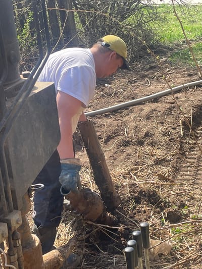 A construction worker in Поедуги, Russia, wearing a yellow cap, white t-shirt, and work gloves, is operating a piece of heavy machinery, possibly for excavation or earthmoving. The worker is holding a muddy log. The environment appears to be a construction site with exposed dirt and scattered debris, including pipes. Vegetation, possibly overgrown, is visible in the background.