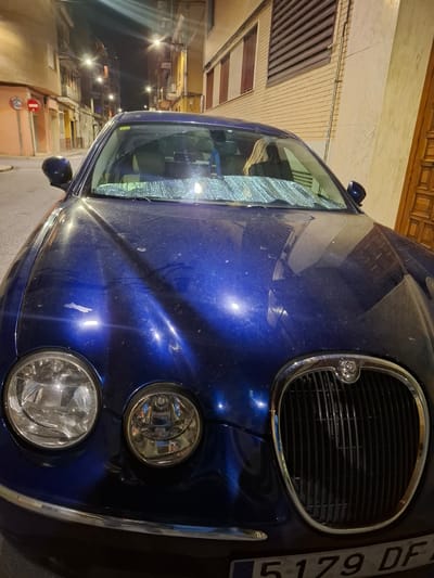 The image captures the front of a blue Jaguar S-Type car parked on a street in Xirivella, Spain. The car has a visible license plate that reads "5179 DF." A sunshade is placed inside the windshield. The street scene shows buildings and streetlights, indicating a nighttime setting in a residential area.