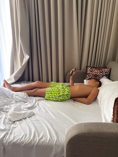 A boy in patterned green shorts lies on his side in a bed with white linens. He faces the curtains and window, holding a phone in his left hand. Part of a grey headboard and accent pillows are visible. The photo is taken from above, with the top of the image showing the head of the bed.