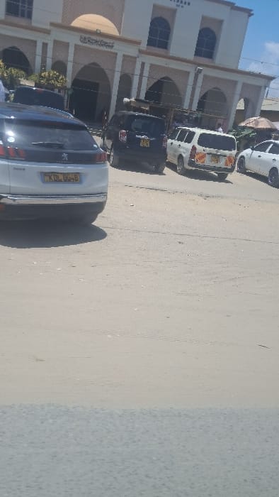 The image displays a daytime street scene in Shimo La Tewa ward, Kenya. A prominent, light-colored building with a central dome, arched windows, and entrances dominates the background. Arabic script is visible above a central archway, and the partial word "JIBAR" is discernible on the upper facade, identifying it as a mosque. The street, composed of a light-colored, dusty ground and a darker paved section in the foreground, is occupied by several vehicles. A white Peugeot 3008 SUV, license plate KDL 004B, is in the immediate foreground. Behind it, to the left, is a dark-colored SUV with a partially visible Kenyan license plate (ending in 964X). To the right of the dark SUV is a white Toyota Probox van with orange and yellow reflective markings and license plate KCG 191T. A white sedan is partially visible further to the right. The sky is hazy blue, indicating clear weather. A street vendor's stall with an umbrella is partially visible on the far right. Specific individuals or their actions are not distinctly discernible.