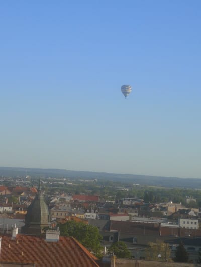 An elevated long shot captures a hot air balloon gracefully ascending over the cityscape of Pardubice, Czechia. The sky is a vast expanse of clear, bright blue, dominating the upper two-thirds of the frame. The hot air balloon, decorated with distinct white and blue patterned stripes, floats in the upper right portion of the sky.
Below the clear sky, the image reveals a sprawling view of the town. Numerous buildings with varied rooflines and colors are visible, many featuring characteristic red-orange terracotta tiles. A prominent, dark-colored domed spire or church tower with a sharp finial stands out in the mid-ground, rising above other structures. Green trees are interspersed throughout the urban landscape, adding patches of natural color.
In the far distance, a gentle range of hills or low mountains forms the horizon, covered in dense greenery. The overall atmosphere is peaceful and serene, with the clear weather and soft lighting suggesting either early morning or late afternoon. No individuals are visibly present, and there is no discernible text in the photograph. The scene depicts a tranquil hot air balloon flight over a picturesque Central European town.