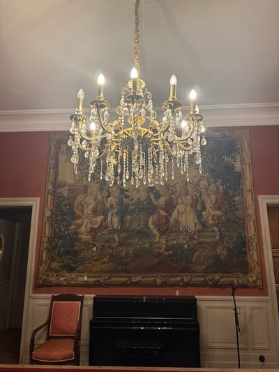 The image depicts an interior scene, likely a formal room, with a prominent ornate golden chandelier hanging from the ceiling. Below the chandelier, a large tapestry hangs on a coral-colored wall, featuring a pastoral scene with figures in classical attire. To the left of the tapestry, an open doorway leads to another room. In front of the tapestry, a black upright piano sits on a white paneled wall. To the left of the piano is a wooden chair with a red patterned seat and back. A microphone stand is positioned to the right of the piano. The lighting suggests an evening or indoor setting. The location is identified as Lège-Cap-Ferret, France.