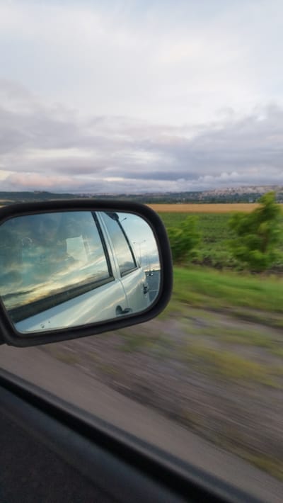 The image captures a view from inside a vehicle moving along a road. In the foreground, the side of the car is visible, and to the left, a side-view mirror reflects the sky, clouds, and part of the car's interior, including a faint silhouette of a person. Beyond the mirror, a blurry landscape unfolds with a green field, trees, and a distant city or town. The sky is overcast with patches of light breaking through, suggesting either dawn, dusk, or a partly cloudy day. The motion blur indicates the vehicle is in transit. The location is identified as Novosolovka, Russia.