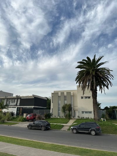 The image shows a street scene in Partido de Quilmes, Argentina. A tall palm tree stands beside a two-story building with a light-colored facade. Two dark-colored vehicles are parked along the side of the street. The sky is filled with white and grey clouds. There are two houses with modern architectural design. There are some small plants at the side of the road.