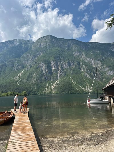 A mountain rises behind a lake, mirroring reflections on the water's surface. A wooden pier juts into the lake, where people stand and a boat is docked. Another boat is adjacent to the pier, sitting on the water's edge. The sky has scattered clouds.