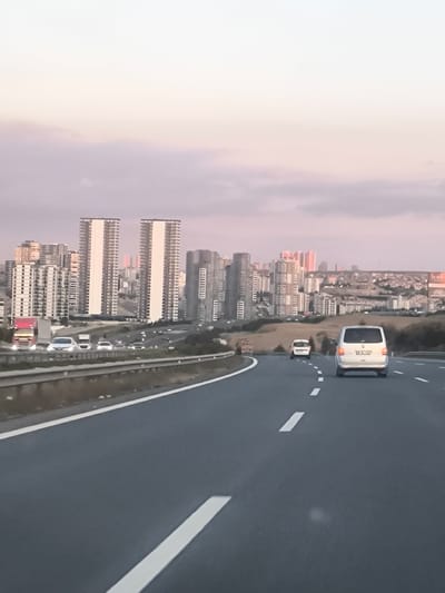 The image depicts a multi-lane highway leading towards a sprawling city skyline under a pastel-colored sky, suggesting late afternoon or early evening. The sky transitions from a pale blue at the top to soft pink and lavender hues towards the horizon.

In the foreground, the dark asphalt road is clearly visible with distinct white lane markings, including both dashed and solid lines. A white van, possibly a Volkswagen Transporter, is seen from behind in the right lane, its license plate partially visible starting with "06". Further ahead in the same lane, another white car drives. In the left lanes, a red truck is visible closer to the camera, with other vehicles receding into the distance. Grey guardrails line the edges of the highway.

The mid-ground is characterized by a dense urban environment featuring numerous high-rise buildings and complexes. Two prominent, tall white buildings with many balconies stand out in the center of the cityscape, appearing almost identical. Other buildings, varying in height and color, populate the area, extending onto distant hills. Some structures with a reddish-orange hue are discernible further back in the skyline. On a distant hill to the right, a faint billboard has visible text that appears to be "bard" or "bird".

The scene captures a typical highway view approaching a large metropolitan area, with the soft, diffused light of twilight lending a calm ambiance to the urban landscape. The setting aligns with Etimesgut, Turkey, a district of Ankara (indicated by the "06" license plate prefix). No people are visible.