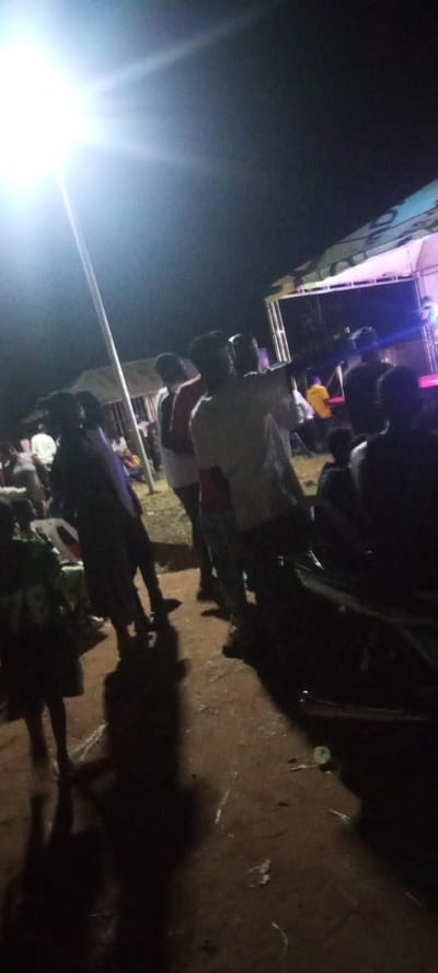 The image captures an outdoor gathering at night, likely a public event or celebration. A bright spotlight illuminates the scene from the upper left, casting long shadows. In the background, a stage or platform is visible, suggesting a performance or presentation is taking place. Several people are gathered, some standing and observing, while others appear to be interacting. The ground is a mix of dirt and what appears to be gravel. The environment suggests a rural or semi-urban setting. This scene is likely located in Ayere, Nigeria, based on the context provided.
