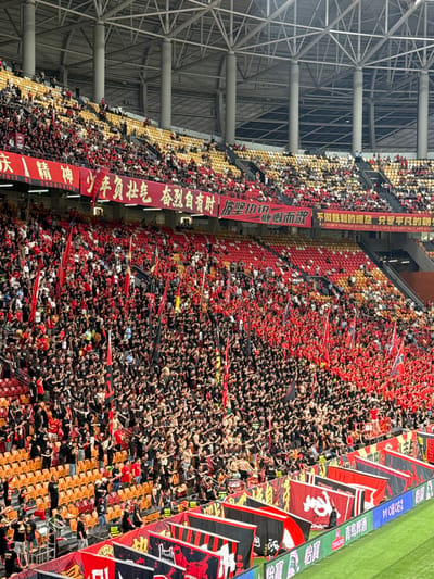 The image shows a dense crowd of people in a stadium, likely in Longxing, China. The crowd is predominantly wearing dark clothing with red accents, holding flags and banners with Asian characters. The stadium seating is a mix of yellow, red, and orange. A large banner with Chinese characters is displayed along the side of the seating. To the left, a metal structure resembling stadium roofing is visible. Adjacent to the field is a blue billboard. The crowd is packed tightly, with many people appearing to be waving or raising their hands.