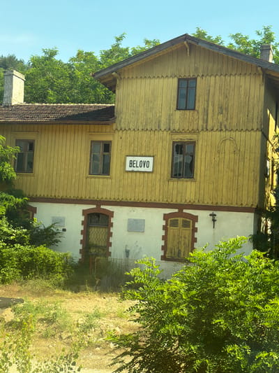 The image shows an exterior view of a weathered, yellow wooden building, identified by a sign as "BELOVO". The building appears to be an old train station, with multiple windows, some of which are broken. The roof is tiled, and a stone chimney is visible on the left side. The building is surrounded by lush green trees and foliage. The sky is clear and blue. The scene suggests a rural or suburban setting, possibly in the town of Belovo, Bulgaria. There are no people visible in the image. The overall impression is one of neglect or abandonment, with the vibrant green vegetation contrasting with the decaying structure.