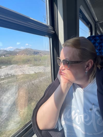 An adult female is depicted inside a train car, gazing out a window at the landscape near Goričani, Montenegro. The subject has light-colored hair, styled in a ponytail, and wears large, rectangular, brown-tinted sunglasses, a light blue V-neck t-shirt with partially visible white text, and a dark, possibly black, zippered jacket. Her right hand supports her chin as she looks outward. Distinctive silver-colored earrings are visible, along with a small, arrow-shaped pin on her left chest. The interior shows a window with a dark frame and a top-mounted shade; reflections are present on the glass. A blue and white patterned train seat is visible to her right. The external view through the window reveals a bright blue sky with minimal cloud cover. The landscape features a wide riverbed filled with light-colored gravel and some water, bordered by green and brown vegetation. In the mid-distance, rolling hills and mountains with sparse plant life are present, along with faint structures in the valley, consistent with the rural area surrounding Goričani, Montenegro. Bright sunlight illuminates the scene.