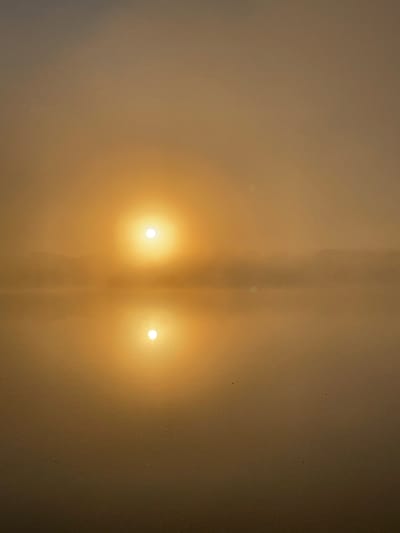 The image shows a foggy sunrise over a body of water, likely a lake or river, with a prominent reflection of the sun. The sky is a hazy golden-orange, and the sun is a bright, diffused orb. The water is still, creating a clear mirror image of the sun and the distant, misty shoreline. Small, dark specks are visible on the water's surface. The overall atmosphere is serene and ethereal. This scene is consistent with a sunrise in the natural areas surrounding Andži, Latvia, characterized by its numerous lakes and misty mornings, especially during certain seasons. There are no people or distinct man-made objects visible in the frame.
