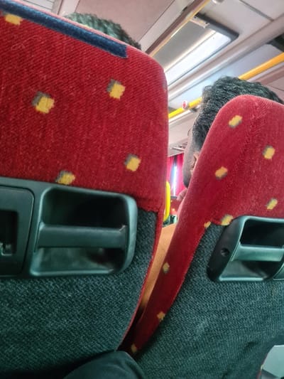 The image displays the interior of a passenger bus in Al-Kiswa, Syria, viewed from a seated position looking forward. Two red and dark grey fabric-upholstered seat backrests, each with a pattern of small yellow squares and a black plastic grab handle, occupy the foreground. The head of a person with dark, curly hair is visible above the left foreground seat. Between and slightly behind the two foreground seats, the head and right shoulder of a second individual, wearing a brown or orange garment and possessing dark, short hair, are visible. Overhead, a rectangular white light fixture and a yellow handrail are present. Red curtains are partially visible through the gap between the seats. A dark fabric object is partially visible in the bottom left corner of the frame.