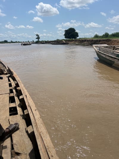 The image depicts a wide, muddy river in Nemnei, Nigeria, under a bright, cloudy sky. In the foreground, the wooden interior of a boat is visible, with a person's left leg wearing a black sandal resting on the wooden planks. The boat appears to be moored or moving slowly.
The river itself is a dominant feature, with its murky brown water reflecting the sunlight. On the far bank, a steep, eroded clay embankment rises, topped with green vegetation and a few scattered trees. The most prominent tree on the far bank is a large, dark-leafed specimen situated on a bend in the river.
In the distance, several other wooden boats are visible, some of them small and flat-bottomed, suggesting local transportation or fishing activities. The overall scene conveys a sense of rural life and the importance of the river as a mode of transit and sustenance in the area. The environment appears natural and undeveloped, with no man-made structures visible other than the boats themselves and possibly some distant, indistinct forms that could be part of the landscape or very distant structures.
