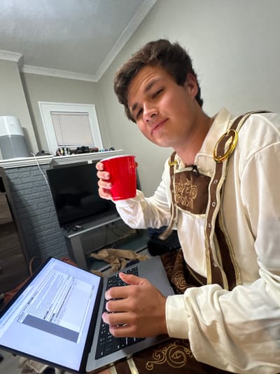 The image depicts a young adult male, presumed to be in a residential interior in South Bend, United States. He has medium-length dark hair and a light complexion, looking directly at the camera with a slight smile. He wears a costume resembling a medieval or fantasy-style tunic, featuring a cream-colored puffy-sleeved shirt with brown and gold-trimmed overlays, secured with two large gold-colored oval buckles. His right hand holds a red plastic party cup, raised approximately to chest height. His left hand is positioned over the keyboard of an open silver laptop (likely a MacBook Pro), which is resting on his lap or a surface below him, displaying text on its screen.
In the background, to the left, a light grey, textured wall feature, possibly a fireplace surround, is visible. Above this feature, a white mantle or shelf holds a grey air purifier on the left and several small, dark objects (possibly electronic controllers or eyewear) clustered near a window on the right. The window features white horizontal blinds. Below the mantle, a dark television screen rests on a silver media console. A crumpled brown paper bag is visible on the floor next to the laptop. The room has light-colored walls and a white ceiling with crown molding. The exact location within South Bend is not discernible from the provided visual information.