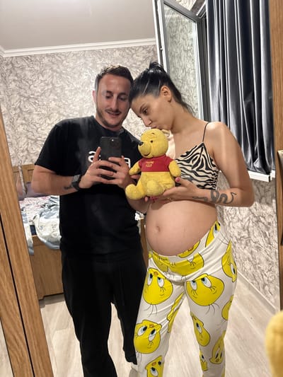 In a mirrored reflection, a man and a pregnant woman are seen in what appears to be a residential interior, possibly in Tbilisi, Georgia. The man, on the left, wears a black t-shirt and black pants, holding a smartphone to take the selfie. He has a short beard and is smiling. The woman, on the right, is pregnant and wears a zebra-print camisole and white pants adorned with cartoon Tweety Bird characters. She is holding a Winnie the Pooh stuffed toy, kissing its head. The background shows a bedroom with a bed and a window covered by gray curtains. The wall is wallpapered with a textured pattern.