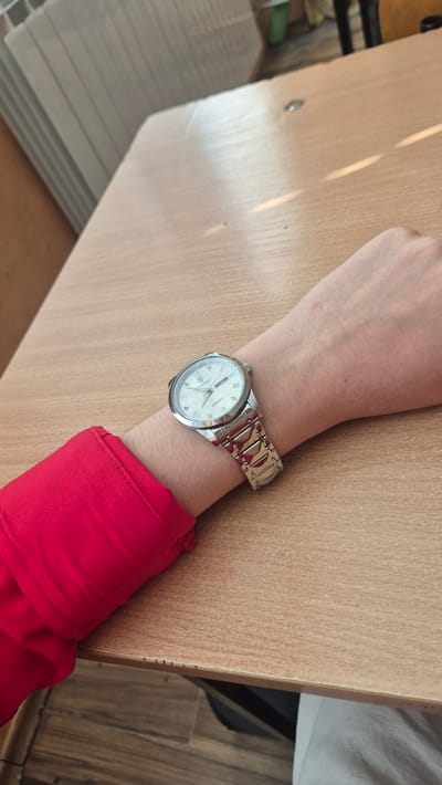 A close-up shot shows a person's left wrist, adorned with a silver wristwatch, resting on a light brown wooden table. The watch face is white with Roman numerals and has a silver band. The person is wearing a bright red long-sleeved shirt. The background is slightly blurred, showing a portion of a white radiator on the left and indistinct objects and flooring on the right. There is no identifiable location in Satovcha, Bulgaria.