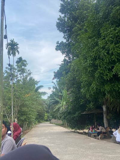 The image depicts a paved road in a rural area of Air Molek, Indonesia. On the left side of the road, several people are seated on chairs, some with their backs to the camera, in what appears to be a roadside seating area. One individual is wearing a red hijab. A tall pole with electrical wires and what appear to be light fixtures is also visible on the left. Lush green trees and vegetation line both sides of the road, with palm trees visible in the distance. On the right side of the road, under a large tree and an umbrella, two people are seated at a small table, with drinks and condiments. A small dog is also present near the table. The sky is partly cloudy. The overall scene suggests a casual, outdoor gathering or resting spot along a rural road.