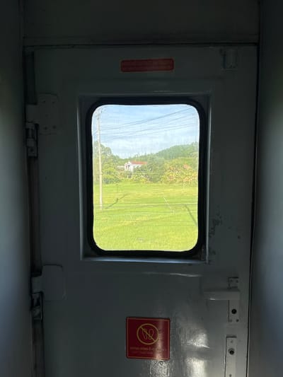 The image shows the interior of a train door with a window looking out onto a rural landscape. The door is painted a muted grey and has a rectangular window with rounded corners. On the door, there are two red warning stickers. The upper sticker, mostly visible, reads in Vietnamese "KHÔNG MỞ CỬA TRƯỚC KHI TÀU DỪNG" and in English "DON'T OPEN THE DOOR BEFORE THE TRAIN STOP". The lower sticker, also red, has a circular graphic with a diagonal line through it and text in Vietnamese and English, which appears to be a "no standing" symbol. Outside the window, a vibrant green rice field stretches towards a line of trees and hills under a bright blue sky with scattered clouds. A utility pole and wires are visible in the field. In the distance, a house with a red roof is nestled among the trees. The scene outside the window suggests the train is moving through the countryside of Bình Chương, Vietnam. The interior of the train is dimly lit, with reflections visible on the door's surface. The door also has a white handle and what appear to be locking mechanisms.
