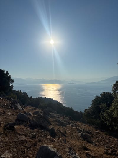 The image depicts a panoramic view of a serene seascape under a clear, bright sky. The sun, a radiant orb, dominates the upper center of the frame, casting a strong, white glare and beams of light that stretch downwards. The sea, a vast expanse of blue, reflects the sun's golden light, creating a shimmering path across its surface. In the distance, a series of hazy mountain ranges form a layered backdrop, their silhouettes fading into the atmospheric perspective. The foreground is a rugged, rocky hillside covered with dry, brown vegetation and scattered shrubs and trees. The terrain slopes downwards towards the sea, with large, grey rocks prominent in the immediate foreground. On the left and right edges of the frame, dark green foliage from trees adds depth and framing to the scene. There are no discernible people or specific structures visible. The overall impression is one of natural beauty and tranquility. This scene could be located in the vicinity of Dalaman, Turkey, which is known for its beautiful coastal landscapes and proximity to mountains.