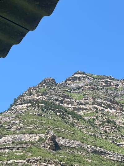 A mountain range with layered rock formations and green vegetation under a clear blue sky. A dark, corrugated object is in the upper left corner, partially obscuring the sky. The mountain slopes are steep and rocky, with patches of grass and scattered trees. The overall scene appears to be a natural landscape under a bright, sunny day.