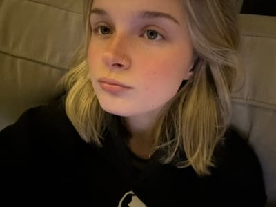 A blonde-haired person lies on a beige surface, possibly a couch. The person wears a dark-colored garment, with a design visible. Their face displays light skin, freckles, and grey eyes. A nose ring is visible. The picture is angled, giving an inverted view of the subject.