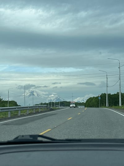 The image shows a view from inside a vehicle driving on a road with snow-capped mountains in the distance. The sky is overcast with thick, grey clouds. Power lines run horizontally across the upper portion of the image. A metal guardrail is visible along the left side of the road. In the middle distance, a white vehicle with red and white signage on its roof is driving in the same direction as the camera. Trees line the right side of the road, and a traffic sign with a blue circle and white arrows is visible. The road is paved and has yellow and white lane markings. The foreground shows the dashboard and windshield of the vehicle from which the photo was taken. The location is identified as the city of Krutoberegovy, Russia.