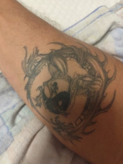 The image shows a tattoo on a person's forearm, against a blurred background of a white and blue striped cloth. The tattoo depicts two stylized dragons intertwined around a Yin and Yang symbol. The dragons are rendered in black ink, with detailed scales and sharp claws. The Yin and Yang symbol is clearly visible in the center of the tattoo. The forearm appears to be lightly tanned and has a few visible hairs. The cloth in the background is soft and textured, suggesting it might be a blanket or a towel. There is no identifiable location in the city of Schwäbisch Gmünd, Germany, visible in the image.