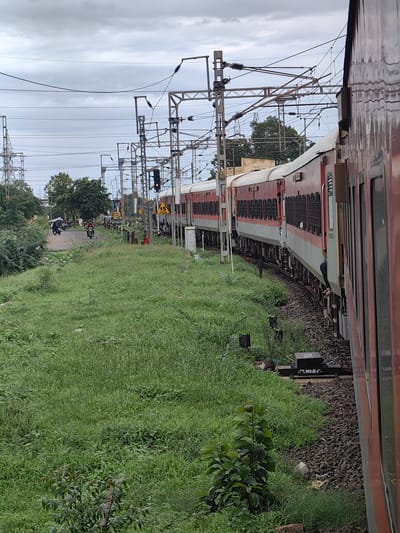 The image depicts a train traveling along a railway track in Kandari, India. The train is red and white with multiple carriages. The track is surrounded by lush green grass and vegetation. Overhead, electrical wires and utility poles are visible against a cloudy sky. In the background, a road with a few motorcycles and people can be seen. There are also several railway signals, including one displaying a red light, indicating that the train may be stopped or preparing to stop. The overall scene suggests an ordinary day at a railway station or along a railway line in India.