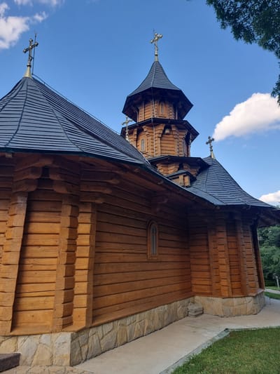 The image shows a low-angle view of a traditional wooden Orthodox church with multiple tiered roofs and spires, each topped with a cross. The church is constructed from logs, with intricate detailing on the eaves and window frames. The main body of the church is visible on the right, with a pathway and grassy area to the right. The sky is blue with some white clouds, suggesting a clear day. The lighting indicates it's daytime. The overall impression is one of serene and rustic architecture. The location is identified as Hagimus, Moldova. No people are visible in the image, and there is no visible text.
