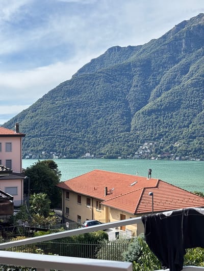 The image displays a panoramic view of Lake Como from Nesso, Italy, under a partly cloudy sky. A large, verdant mountain, densely covered in deciduous trees, dominates the background, rising steeply from the turquoise-colored lake water. Along the distant shoreline on the opposite side of the lake, numerous white and light-colored buildings are visible at the mountain's base.
In the midground, a multi-story building with a terracotta-tiled roof and pale yellow walls occupies the center-right, featuring multiple windows with closed brown shutters. A small, dark rectangular object, possibly a solar panel or vent, is positioned on its roof. To the left, a partially obscured pink-hued building with a similar roof is visible. Various green trees and foliage, including a prominent palm tree, are interspersed between and around the buildings. A dark-colored hatchback vehicle is parked on a paved surface or road behind a dark green metal fence.
The scene is framed from a balcony, indicated by a white railing extending across the bottom of the image. Black clothing, specifically a dark t-shirt or similar garment, hangs drying on a rack in the lower right foreground. No individuals are present in the image.