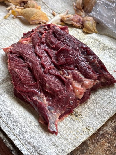 A close-up, overhead shot captures a large, raw cut of dark red beef positioned centrally on a light-colored, textured surface. The beef is thick and irregularly shaped, displaying visible muscle fibers and streaks of fat. The surface beneath the meat appears to be a woven material, possibly a mat or cloth, with a rough texture and a light beige color. In the upper background, out of focus, are several partially prepared raw chickens, some with their legs and feet visible, suggesting a marketplace or butcher setting. A crumpled clear plastic bag is also partially visible in the upper right background. The lighting is bright, creating some shine on the surface of the raw meat. The image was taken on October 15, 2023, at 10:04 AM.