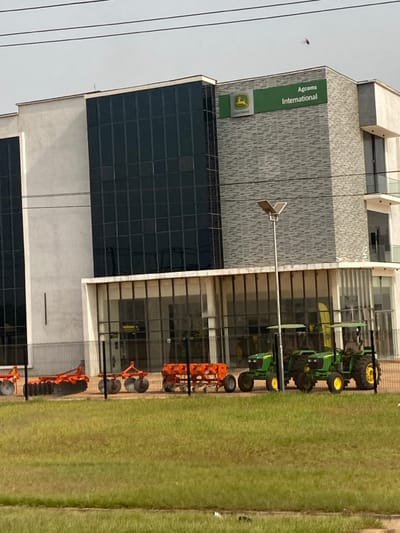 The image displays the Agcoms International building, a John Deere dealership, situated in Issele Uku, Nigeria. The multi-story structure exhibits a modern architectural style, featuring extensive dark glass window panels on its upper levels and light grey, textured wall cladding. A green sign bearing the John Deere logo and "Agcoms International" is mounted on the building's facade. The ground floor is characterized by large glass windows, consistent with a showroom entrance.
A paved display area in front of the building is separated from the immediate foreground by a black metal fence and a strip of green lawn. Positioned on this paved area are multiple pieces of agricultural machinery. These include three distinct orange implements: a disc harrow, a multi-wheeled planter, and an additional tillage tool. Alongside these are two green John Deere tractors, identifiable by their yellow wheels and green chassis. A tall street light, topped with a solar panel, stands between the lawn and the machinery display. The scene is observed under natural daylight; power lines traverse the upper portion of the frame. No individuals are visible.
