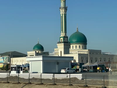 A mosque with multiple green domes and a tall minaret is visible in the background. In the foreground, there are several market stalls with tents set up. A white car is parked near one of the stalls, and a person is riding a bicycle in front of the stalls. A truck filled with watermelons is also parked nearby. The sky is clear and blue. The scene appears to be a bustling marketplace in the city of Baxt, Uzbekistan, with the mosque serving as a prominent landmark.